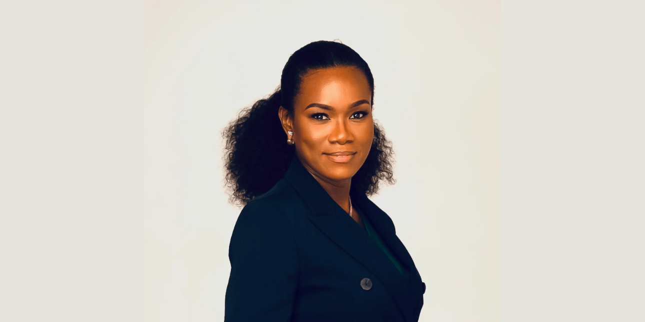 A portrait of a smiling Ivy Heward-Mills. Ivy is a Black woman wearing a dark top with tied back shoulder length hair